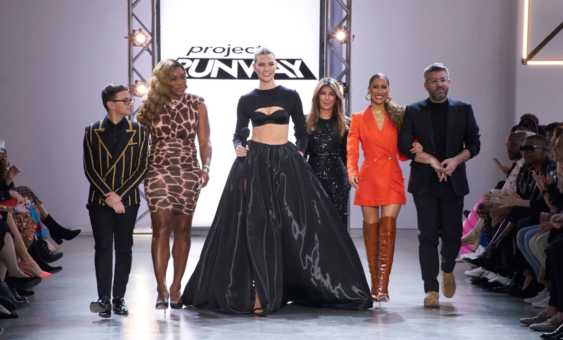 Five people on a runway; three women in dresses and heels, one woman in a crop top and skirt, and a man in a suit. They are smiling