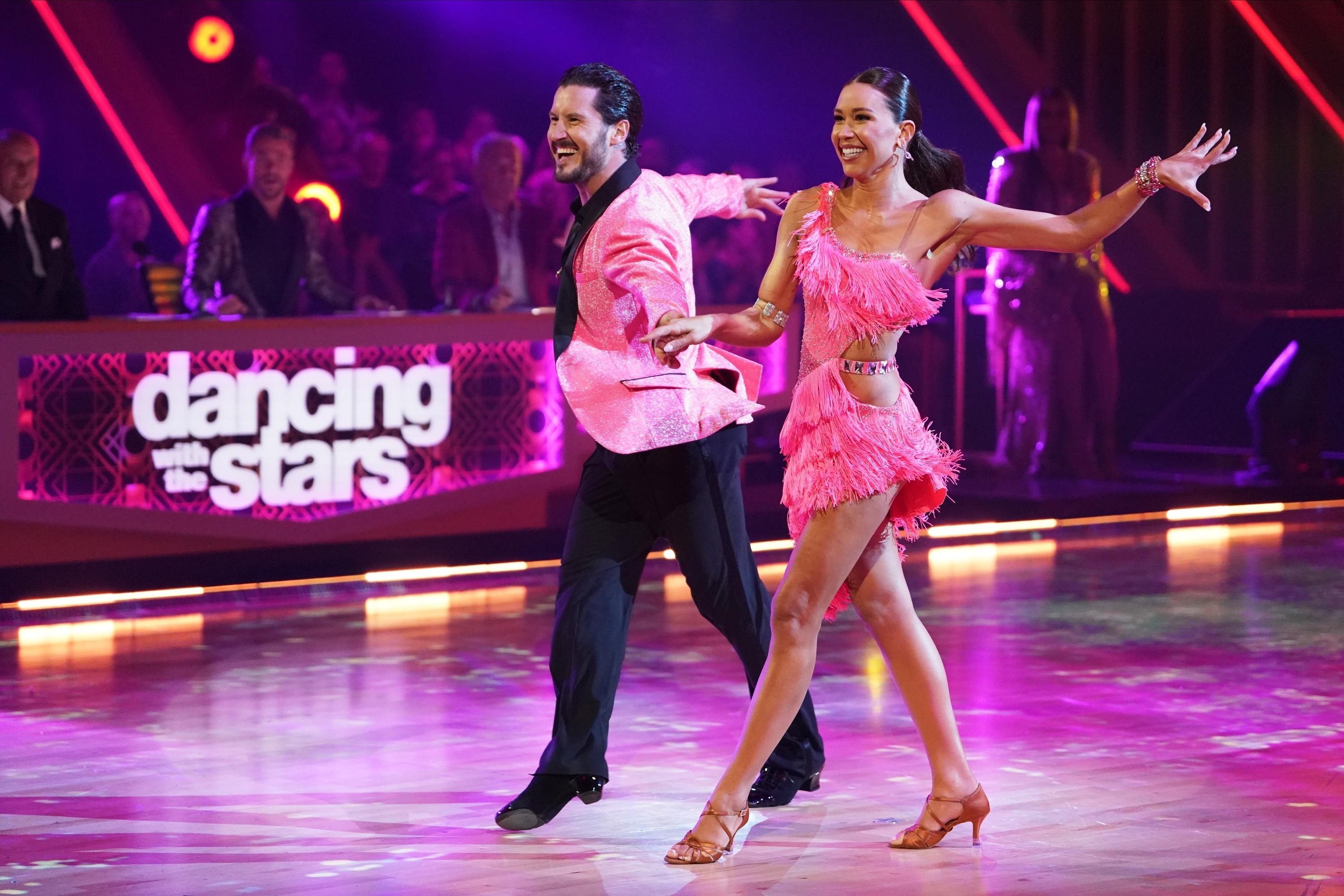 Two dancers perform on stage with smiles, man in a satin jacket and woman in a fringed outfit. The &quot;Dancing with the Stars&quot; logo is visible