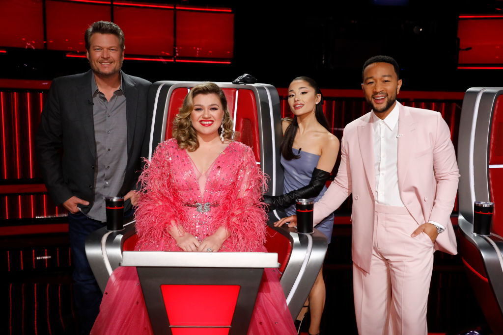 Four coaches from The Voice posing together; two men in suits, two women in dresses, standing by red chairs