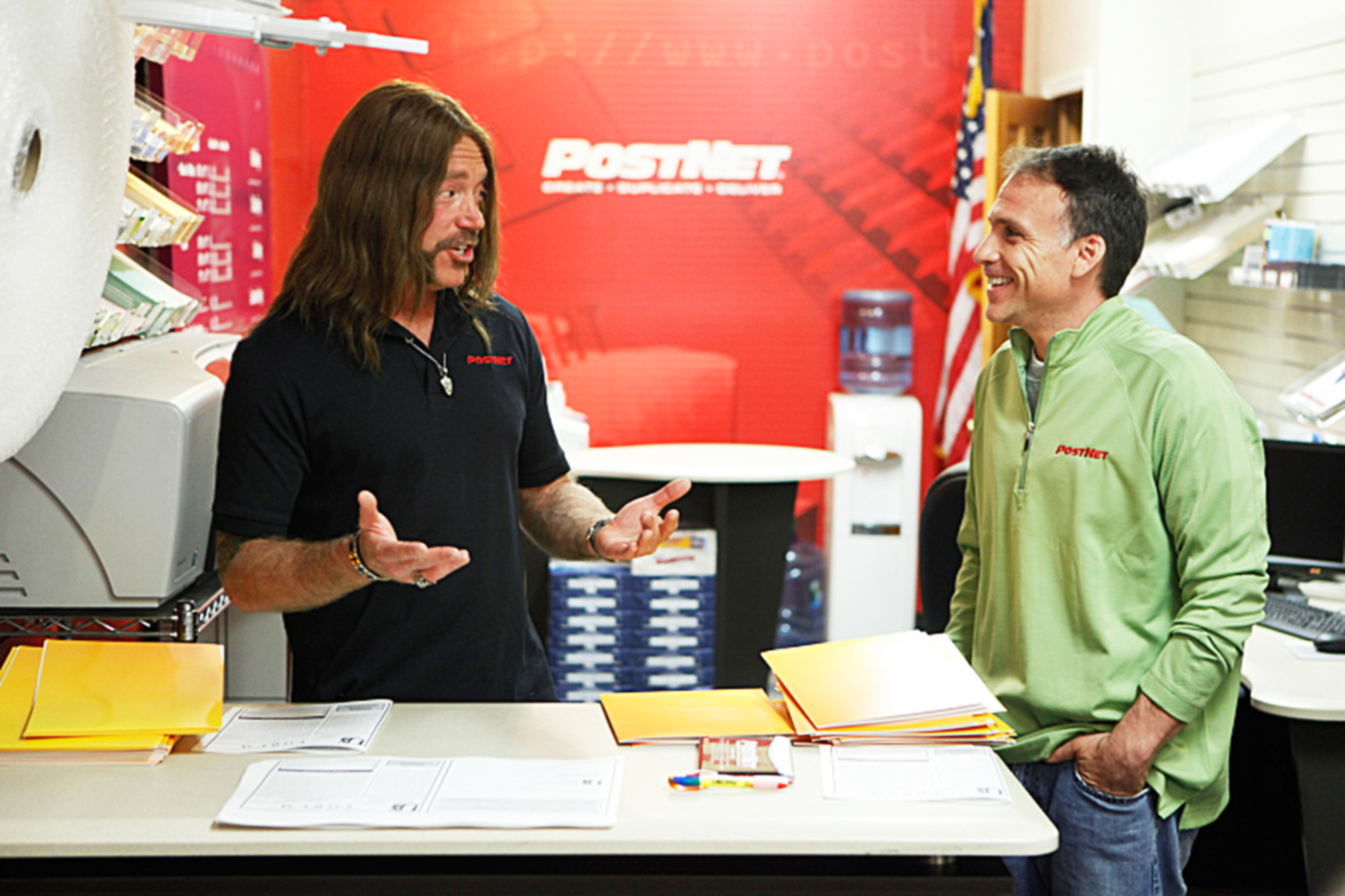 Two people conversing in a store with one person gesturing and the other smiling. Both are wearing branded work attire