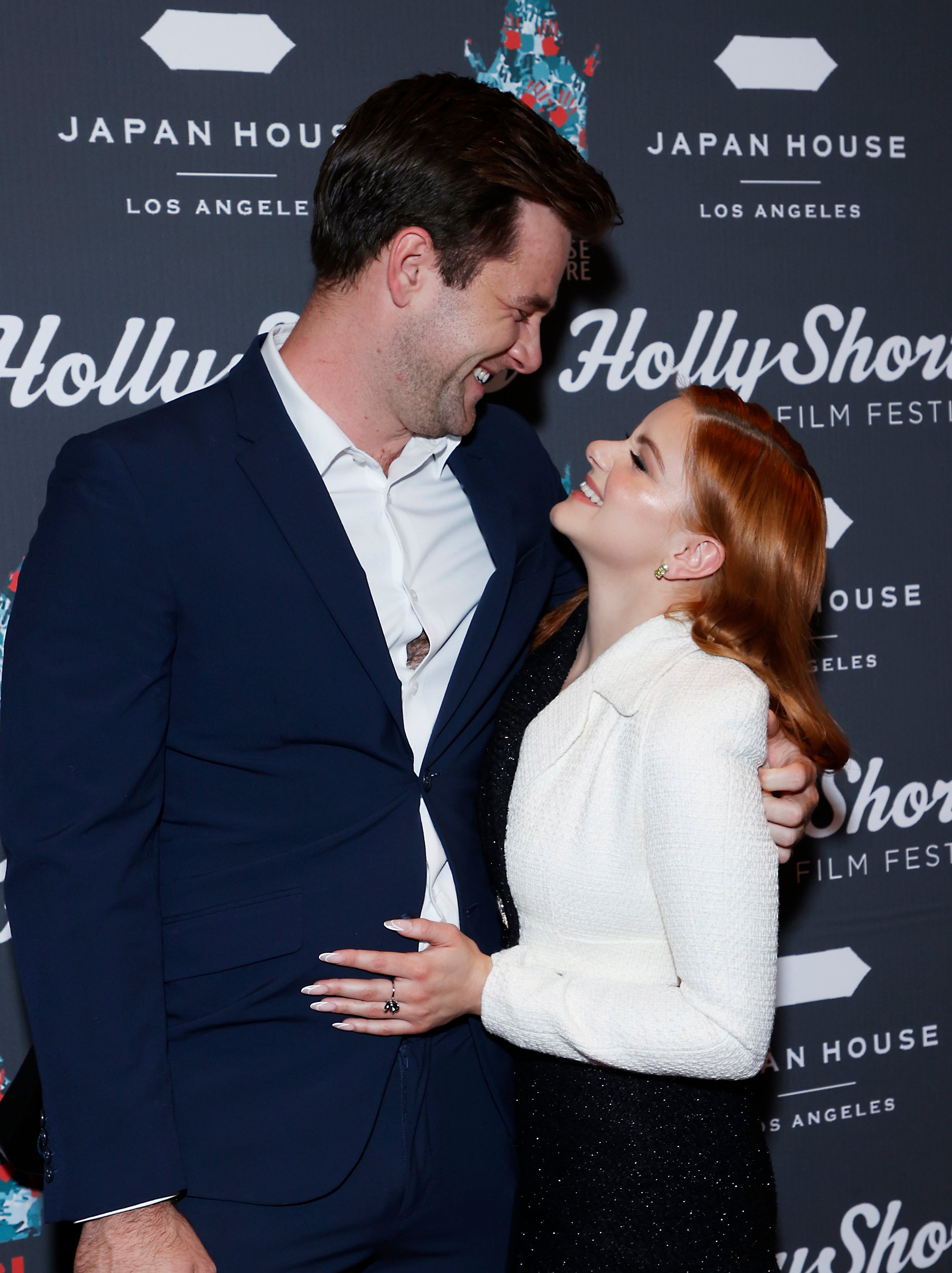 Two people smiling at each other on red carpet; man in a suit and woman in a blazer and shorts