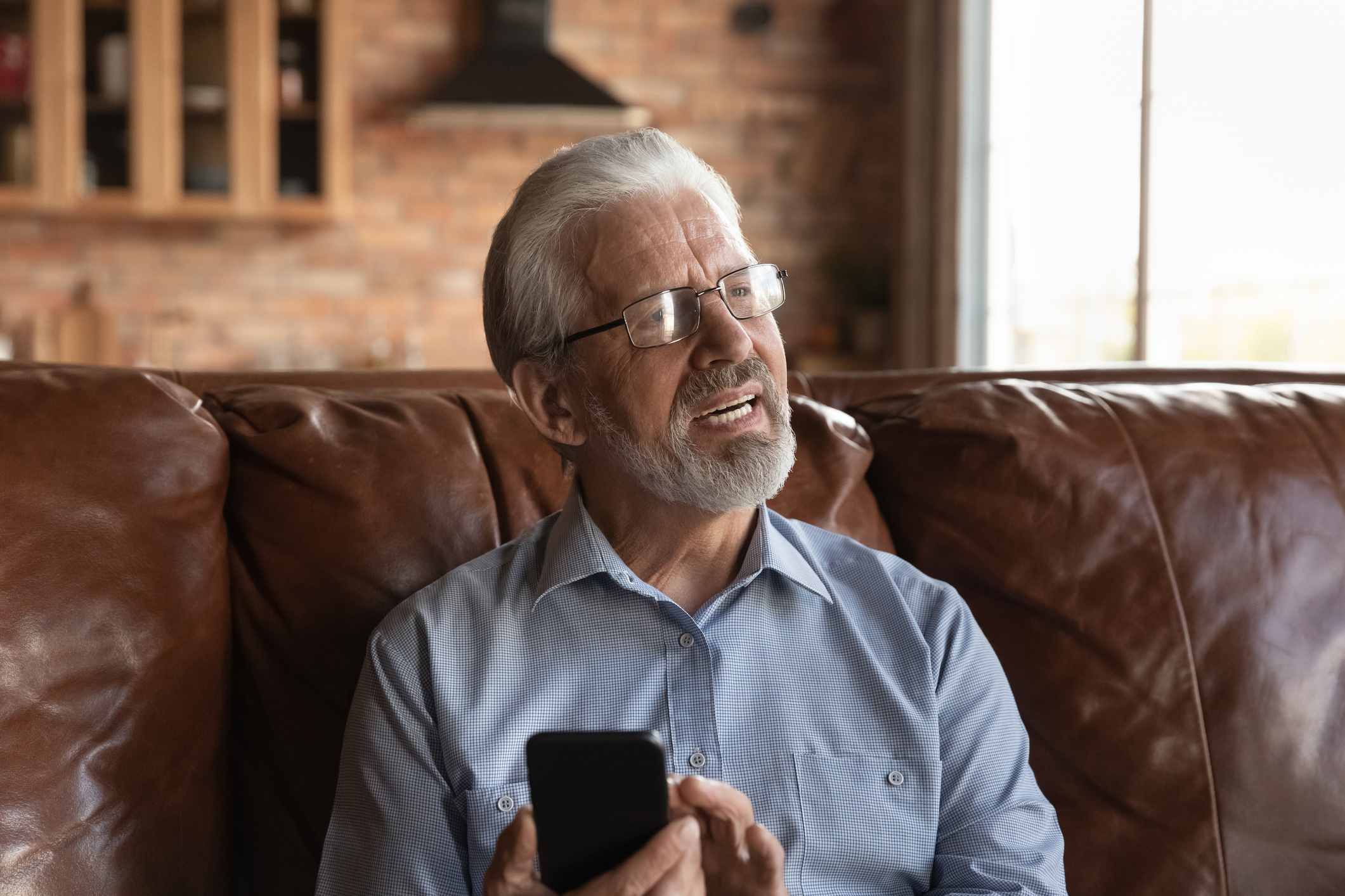 Senior man sitting on a couch looking at a smartphone, displaying a moment of learning or managing technology