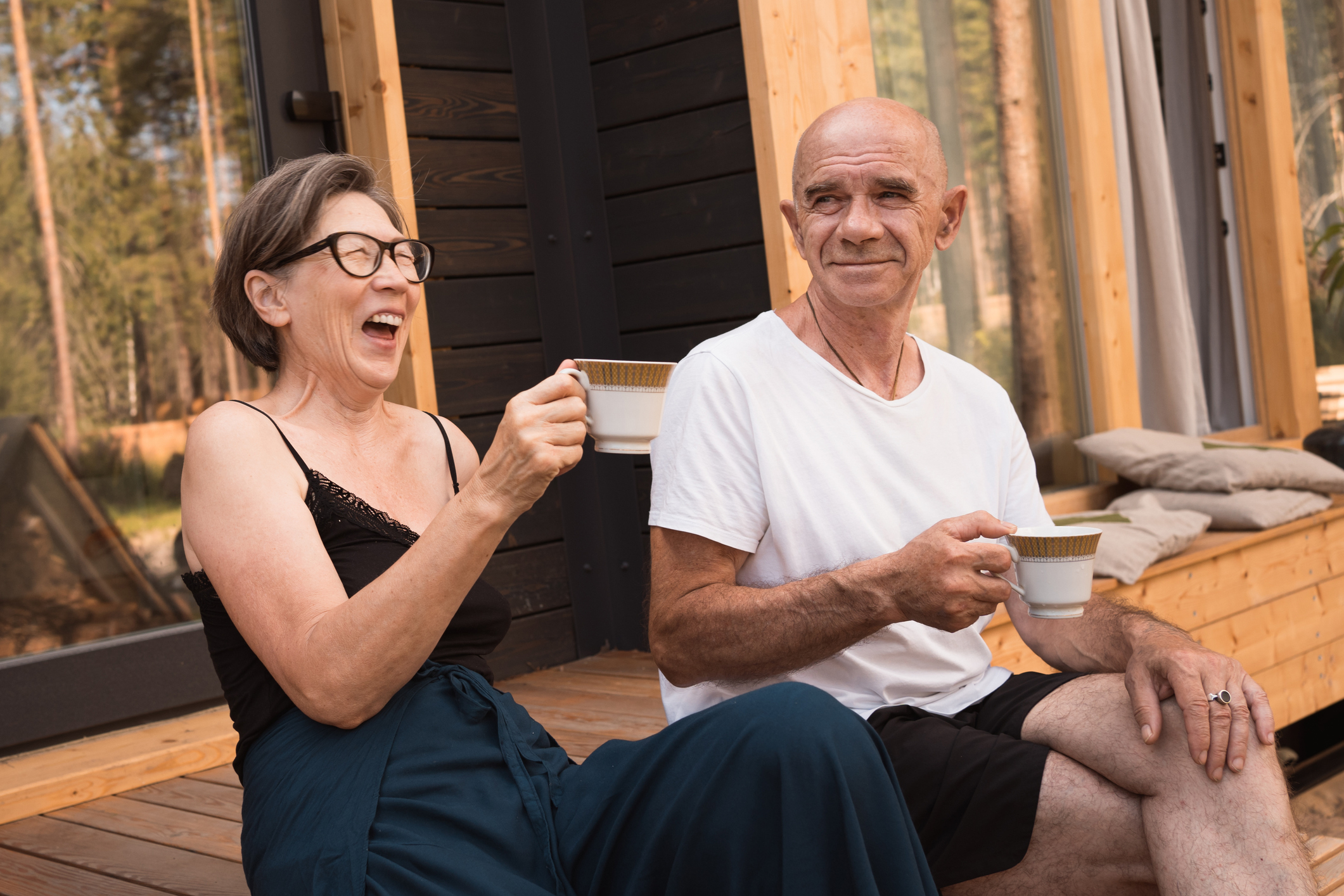Two older adults sharing a laugh while sitting with coffee on a house deck