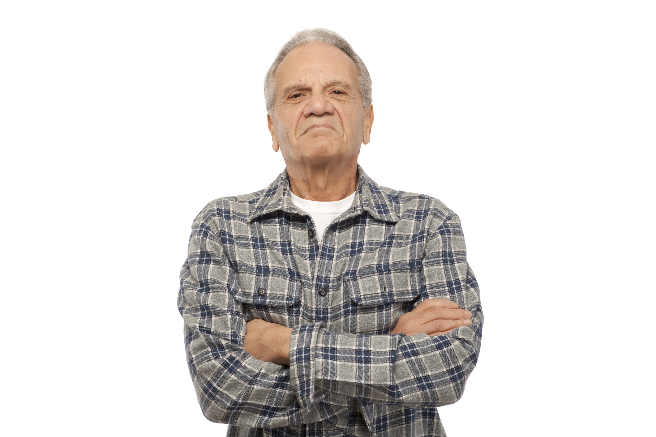 Elderly man with arms crossed wearing a plaid shirt over a white tee, expressing slight discontent.
