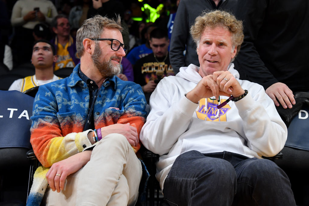 Two men sitting courtside at a basketball game engaged in conversation. One wears a colorful hoodie, the other a white hoodie