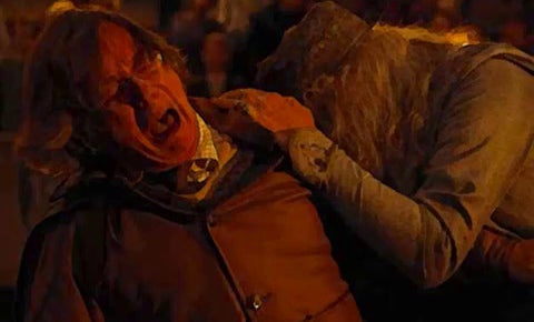 An intense scene from a movie showing a character in period attire being attacked by another in dirty, tattered robes