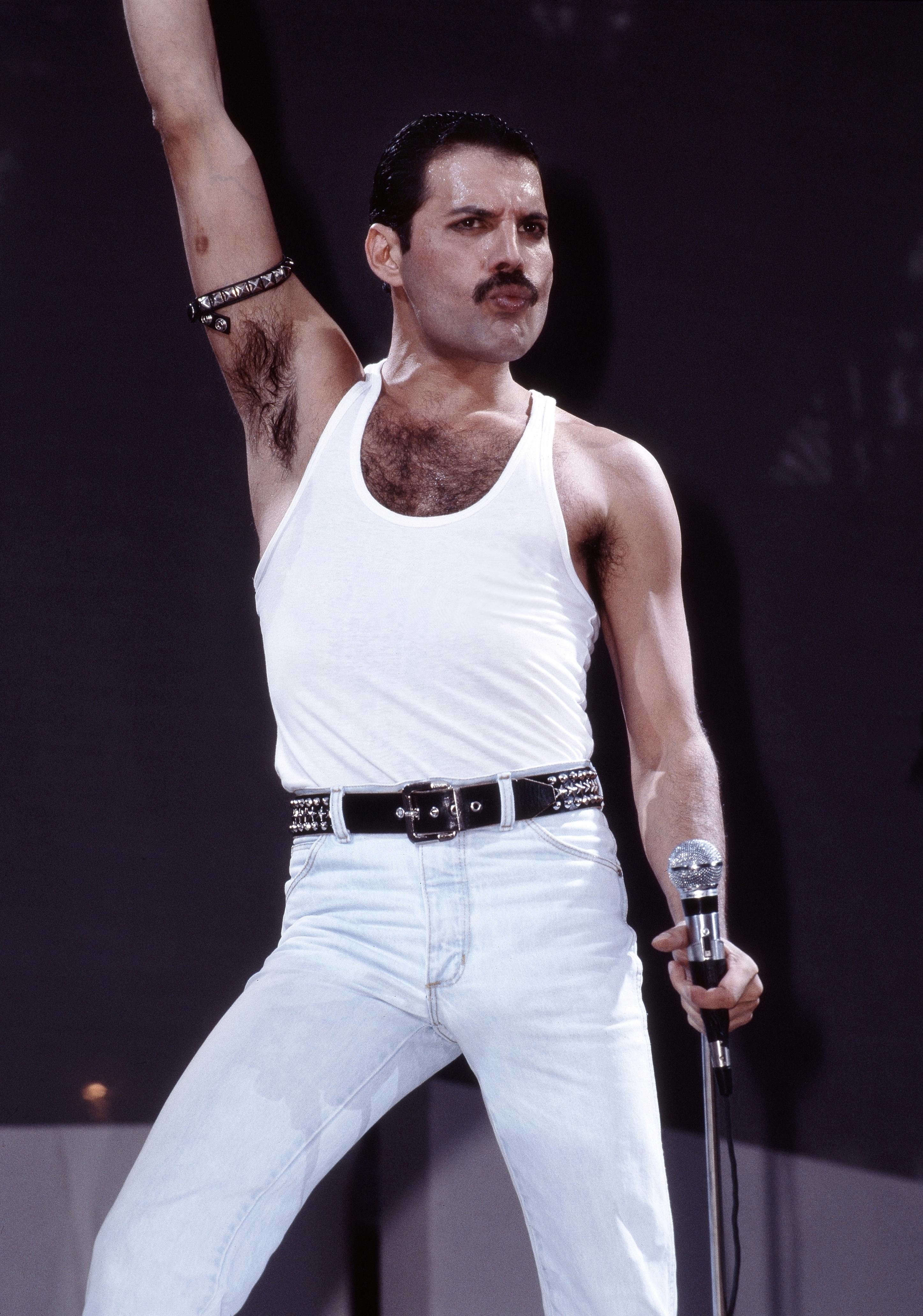 Freddie Mercury on stage in a white tank top and jeans, with one arm raised holding a mic