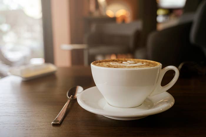A cup of cappuccino with artful foam design on a saucer, spoon to the side, on a table in a cozy cafe setting