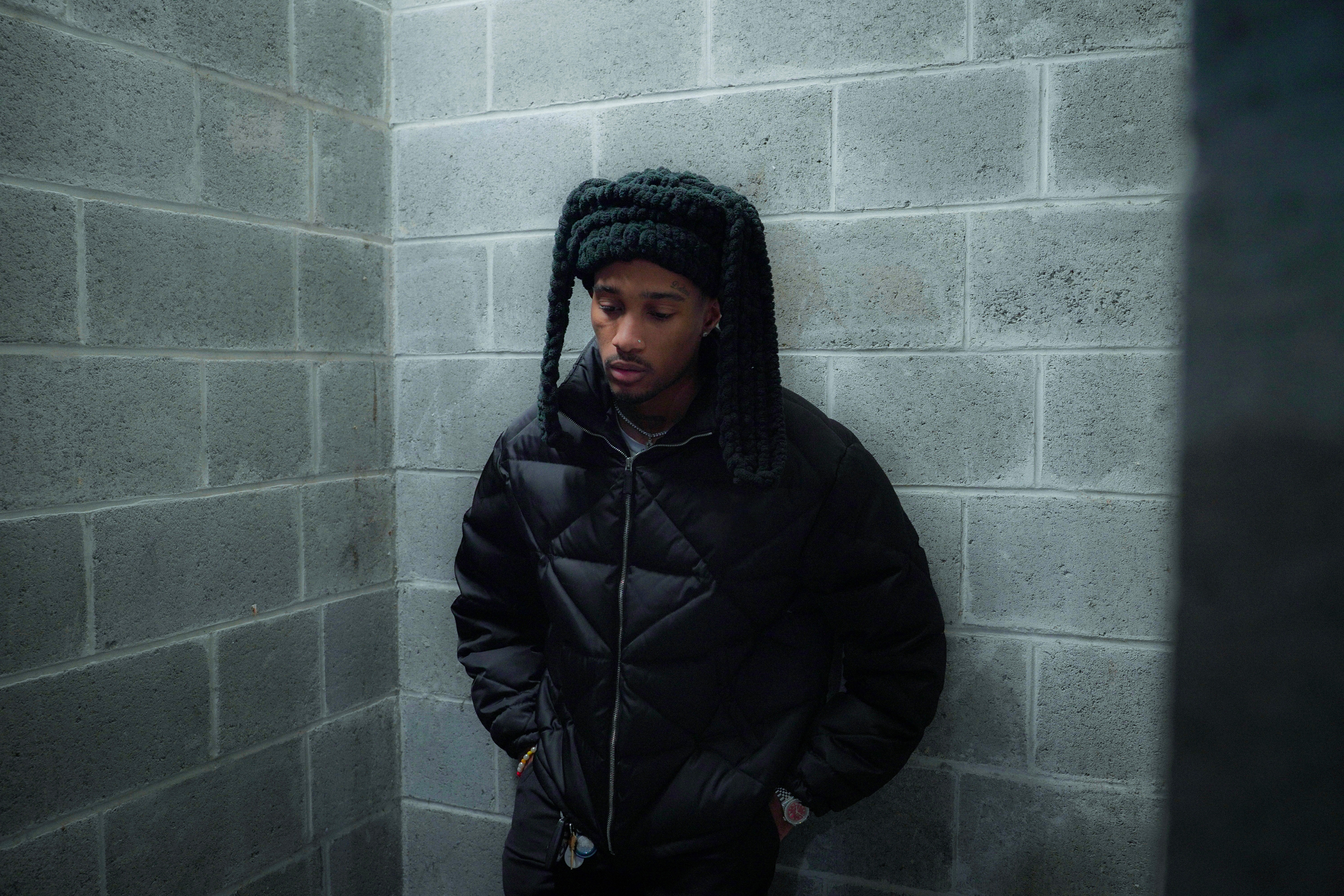 Man in a knitted hat and puffy jacket standing against a cinder block wall, looking thoughtful