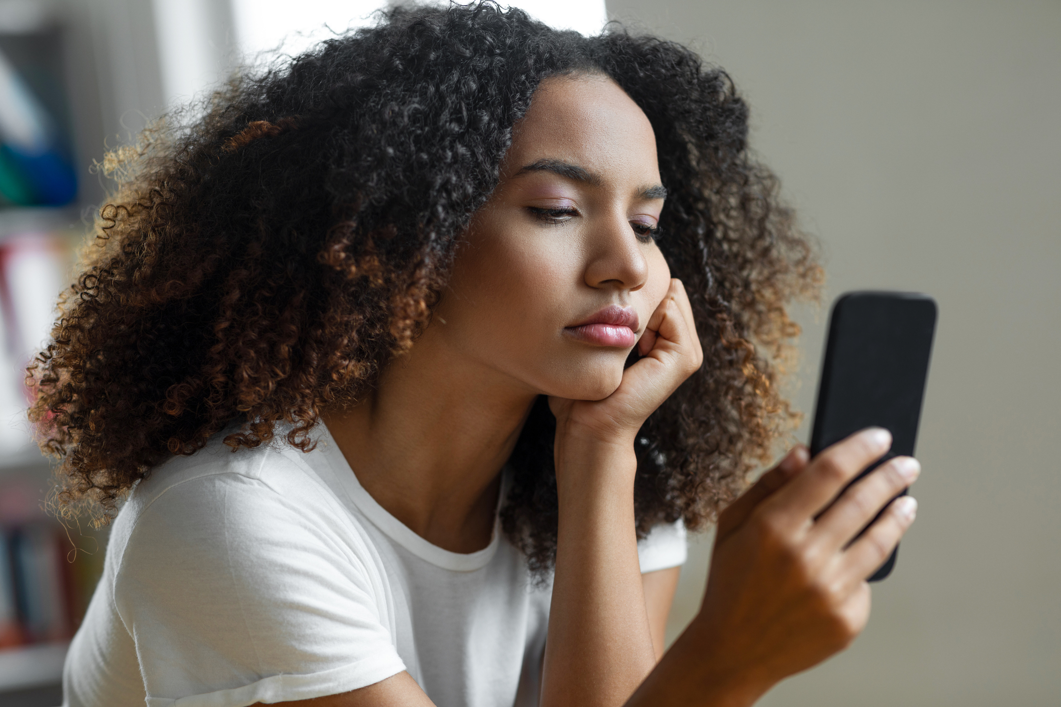 Woman in a white shirt looks at her smartphone, her expression thoughtful or concerned