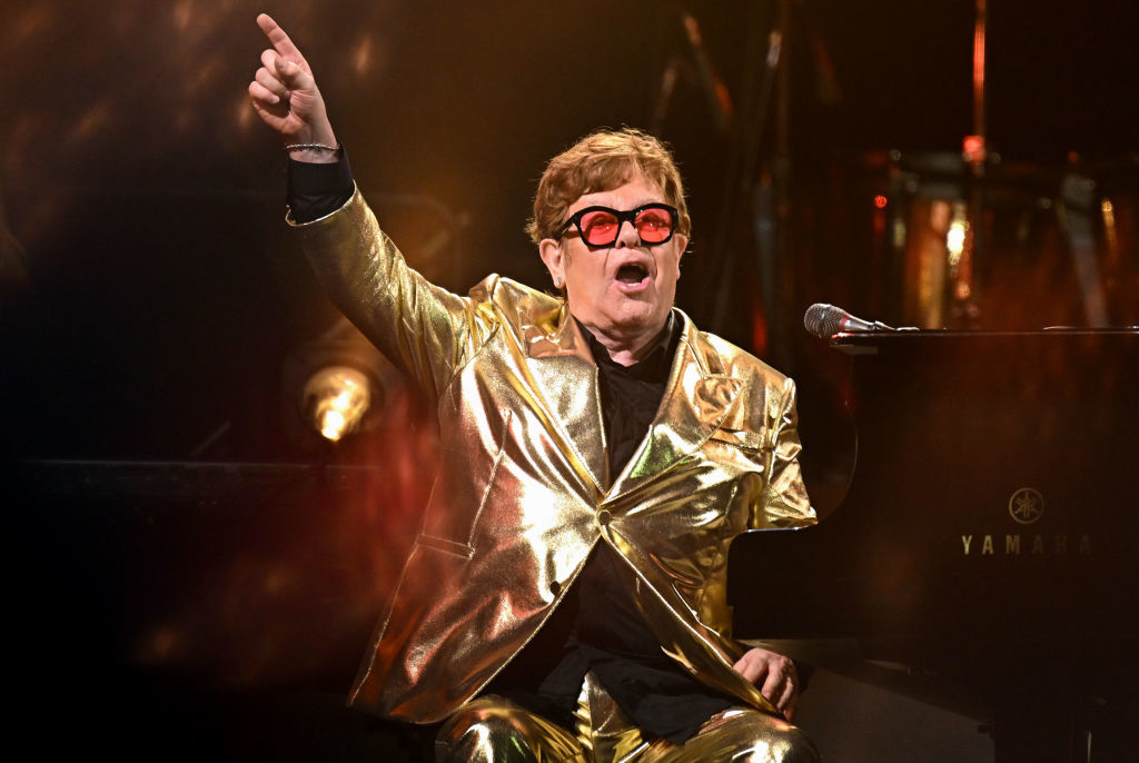 Elton John performing in a gold suit and red glasses, gesturing with one hand up