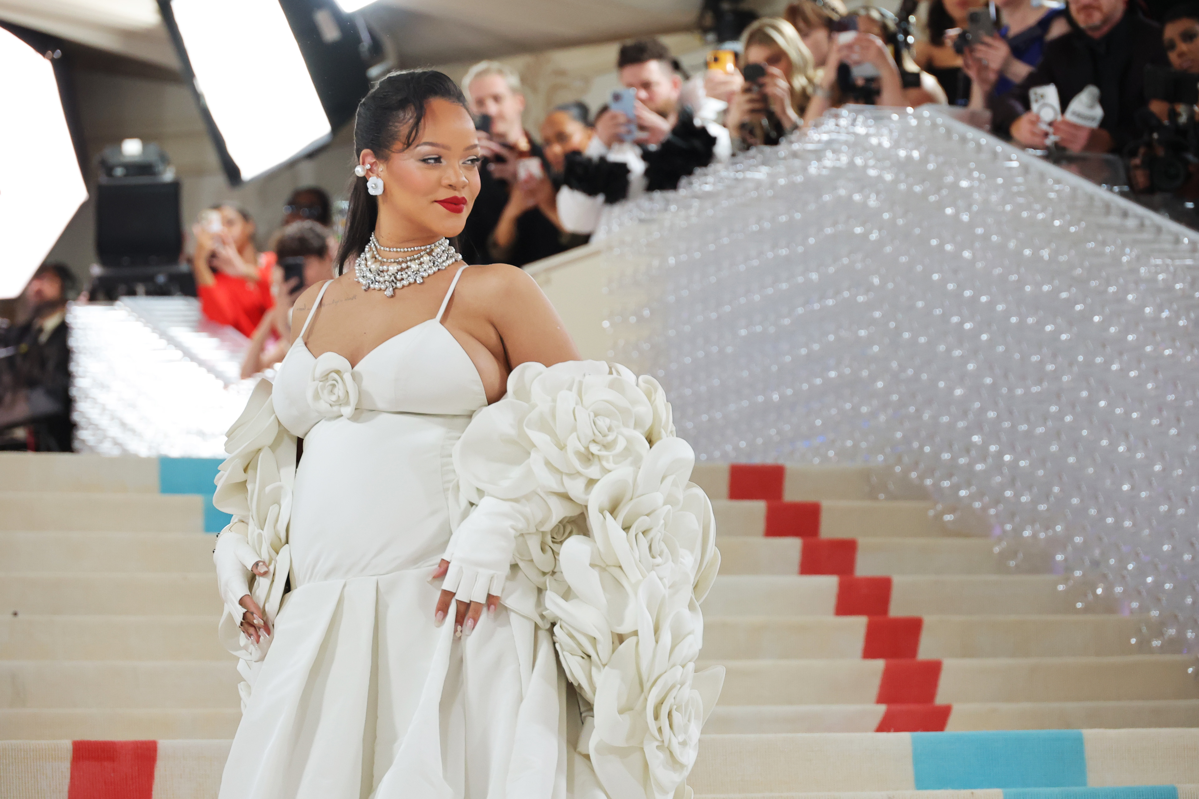 Rihanna in an elegant white gown with large ruffled detail, standing on red carpet steps, photographers in background