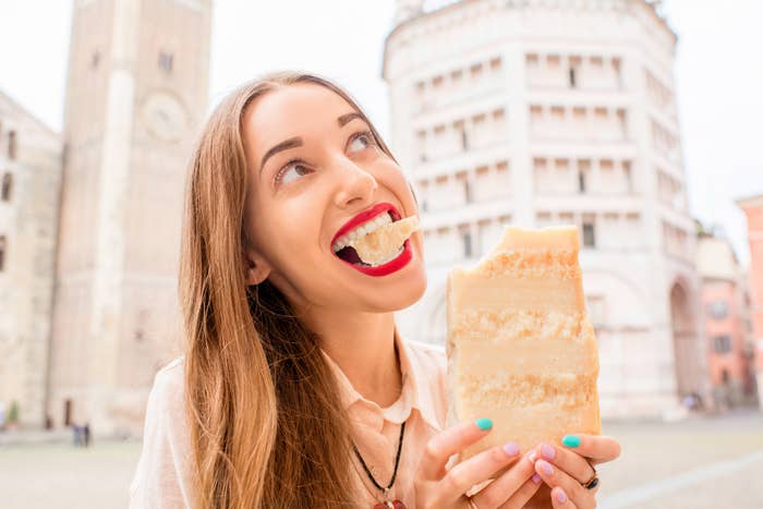 A person smiles while playfully biting into a large piece of Parmesan cheese, standing in front of the Leaning Tower of Pisa