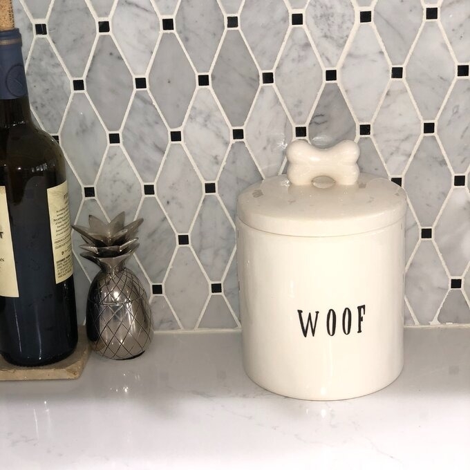 Ceramic dog treat jar labeled &quot;WOOF&quot; with a bone-shaped handle, next to a decorative pineapple. Ideal for stylish pet owners