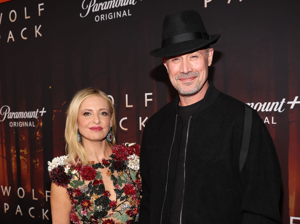 Two people posing at an event; one wearing a floral dress, the other in a black outfit with a hat