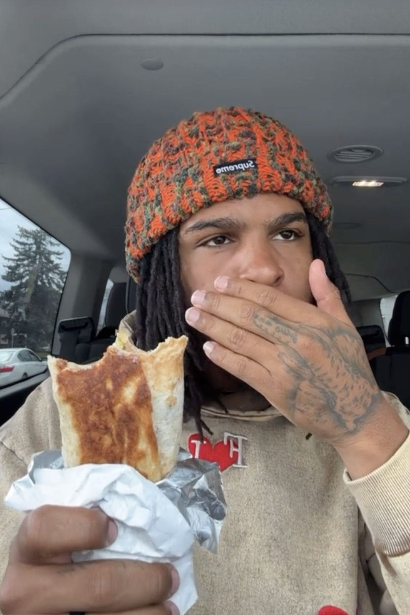 Keith Lee eating a burrito in a car, wearing a beanie and sweater, with visible hand tattoos