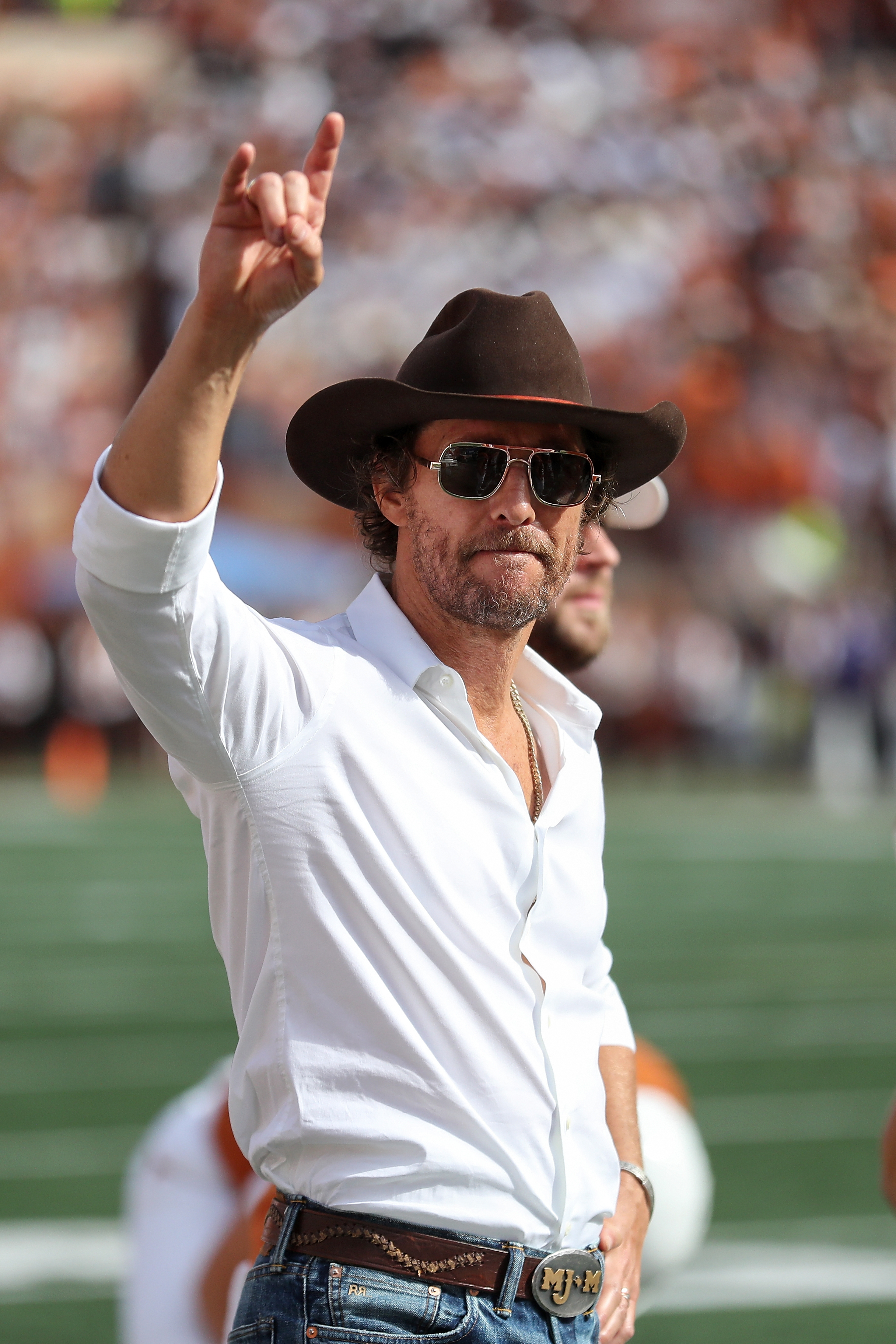 Matthew McConaughey in a white shirt and cowboy hat raising his right hand with two fingers up in a peace sign