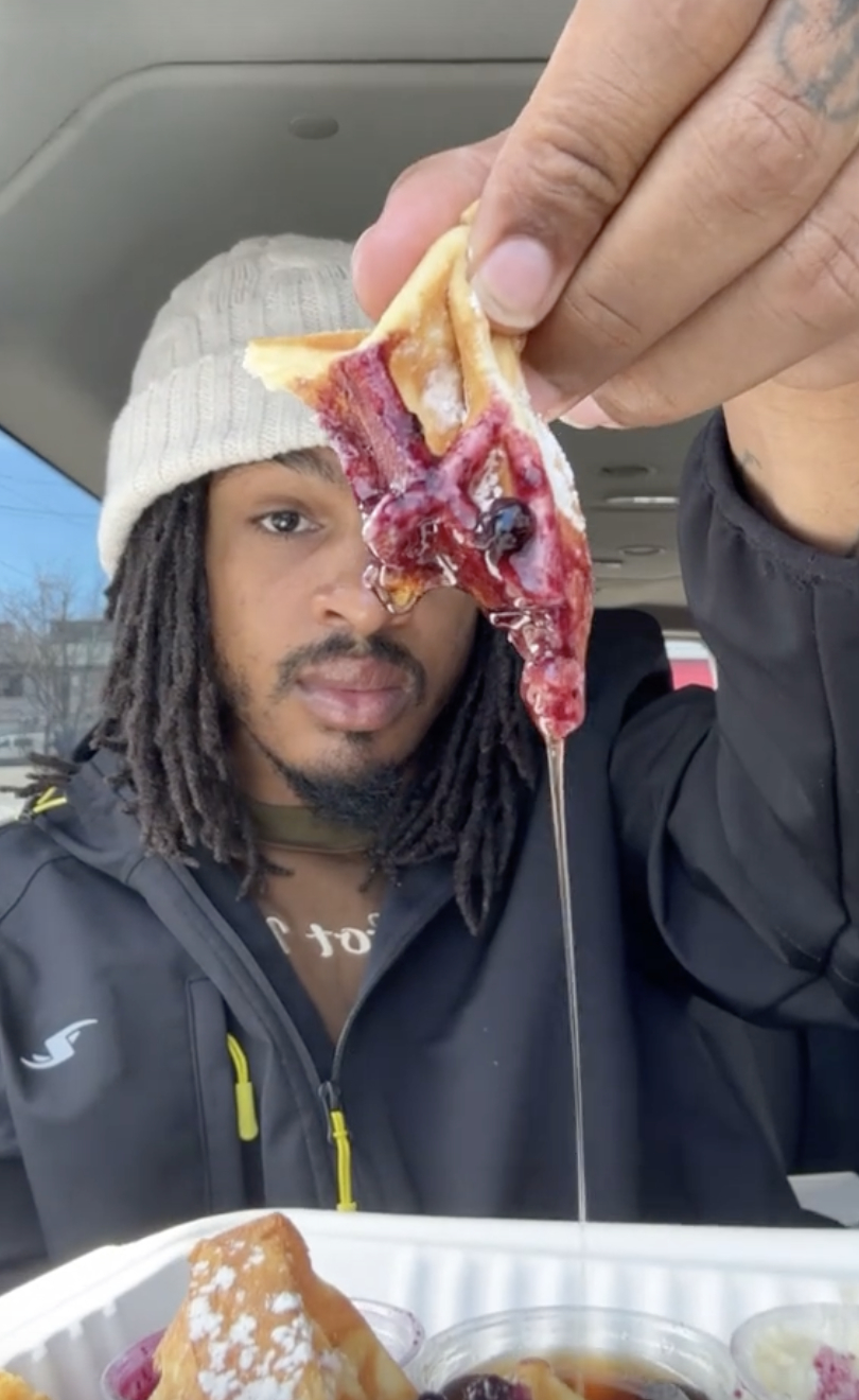 Keith Lee holding a waffle with jam and syrup dripping, wearing a beanie and a jacket