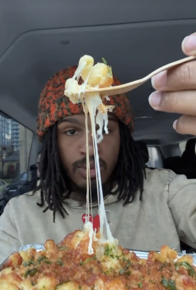 Keith Lee in car holds up a forkful of cheesy pasta, with a full dish visible below