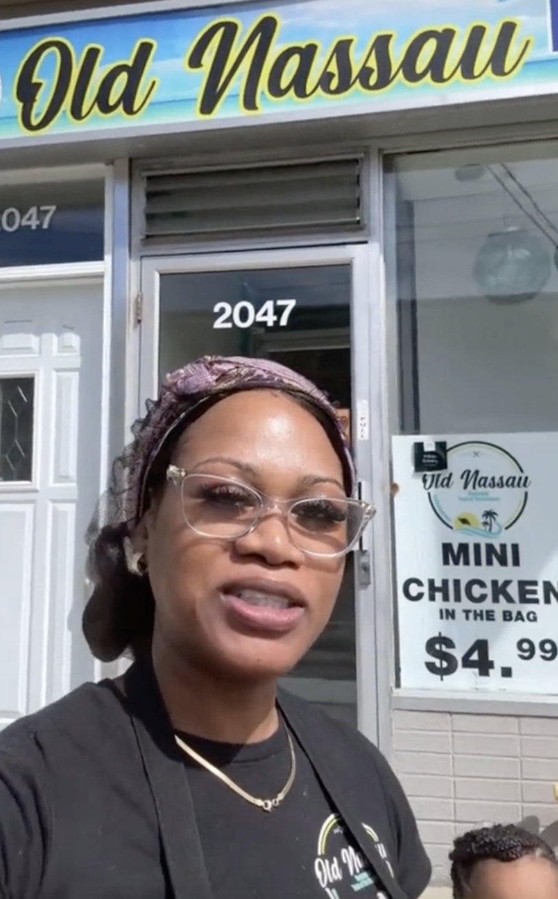 Woman in front of &quot;Old Nassau&quot; store, wearing glasses and headscarf, with &quot;Mini Chicken In The Bag&quot; sign below