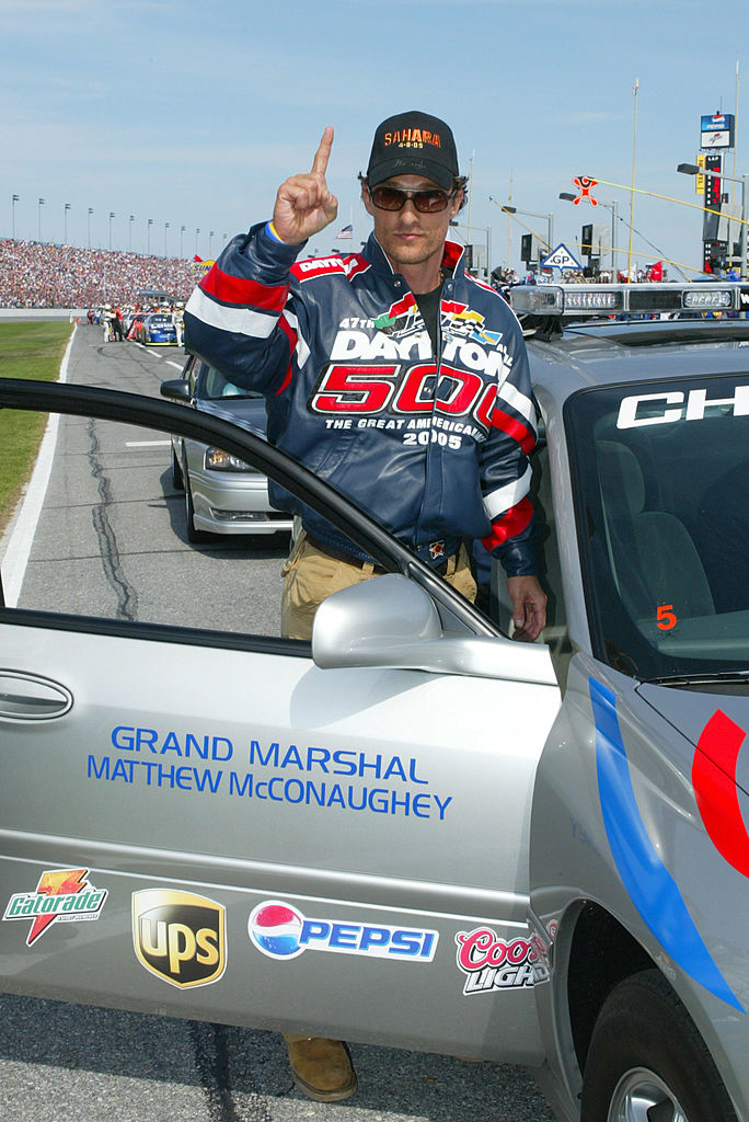 Matthew McConaughey as Grand Marshal at a racing event, standing by a car, gesturing number one with his finger