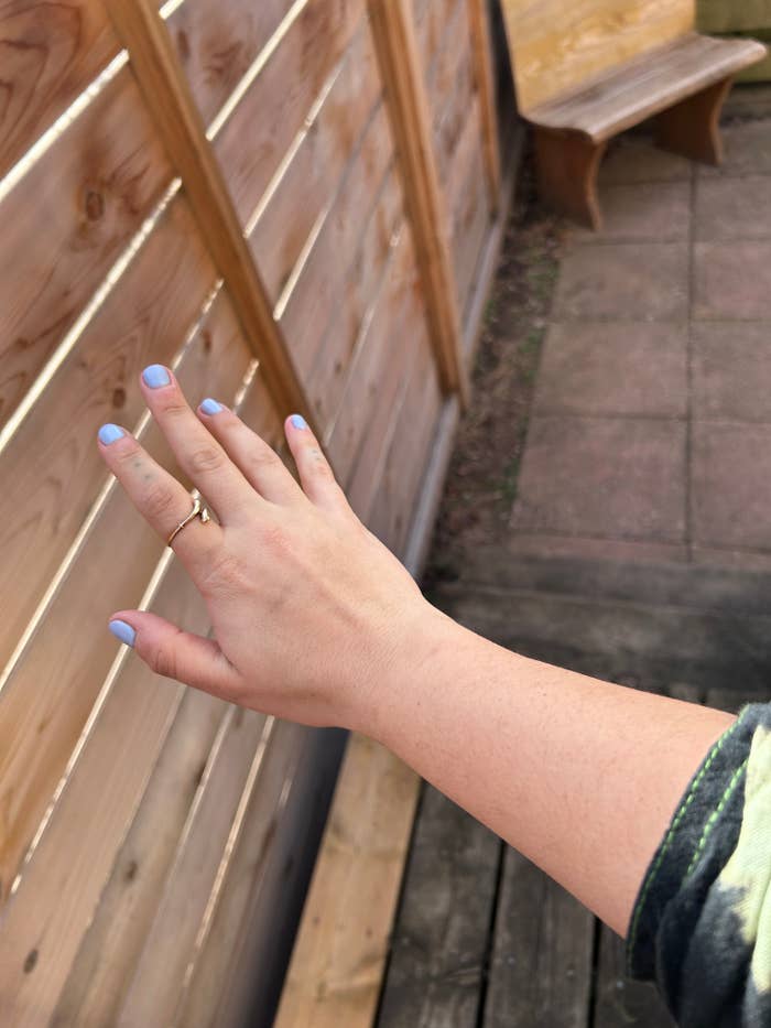 Person's hand with blue nail polish displaying a ring, reaching towards wooden railing outdoors