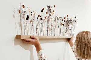 Person hanging a wooden shelf with dried flowers on a white wall, demonstrating home decoration