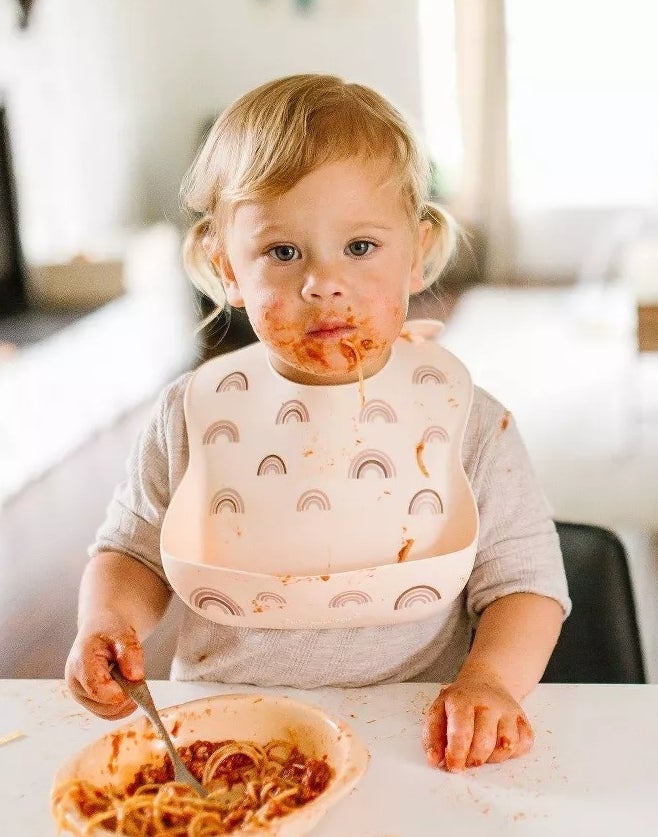 Toddler with spaghetti on face and bib, sitting at a table with a plate of pasta