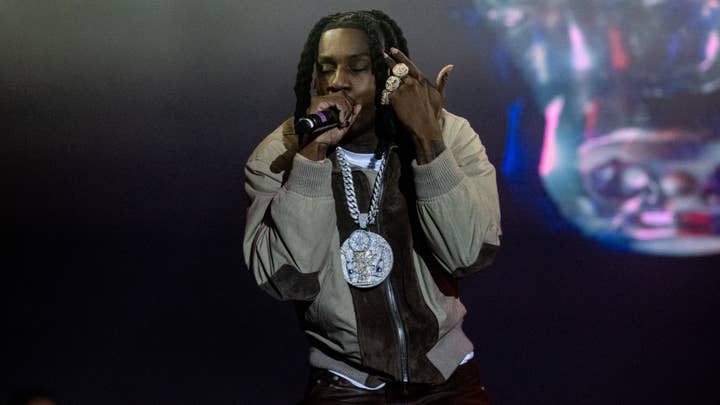 Rapper on stage performing with a microphone, wearing layered necklaces and a casual outfit