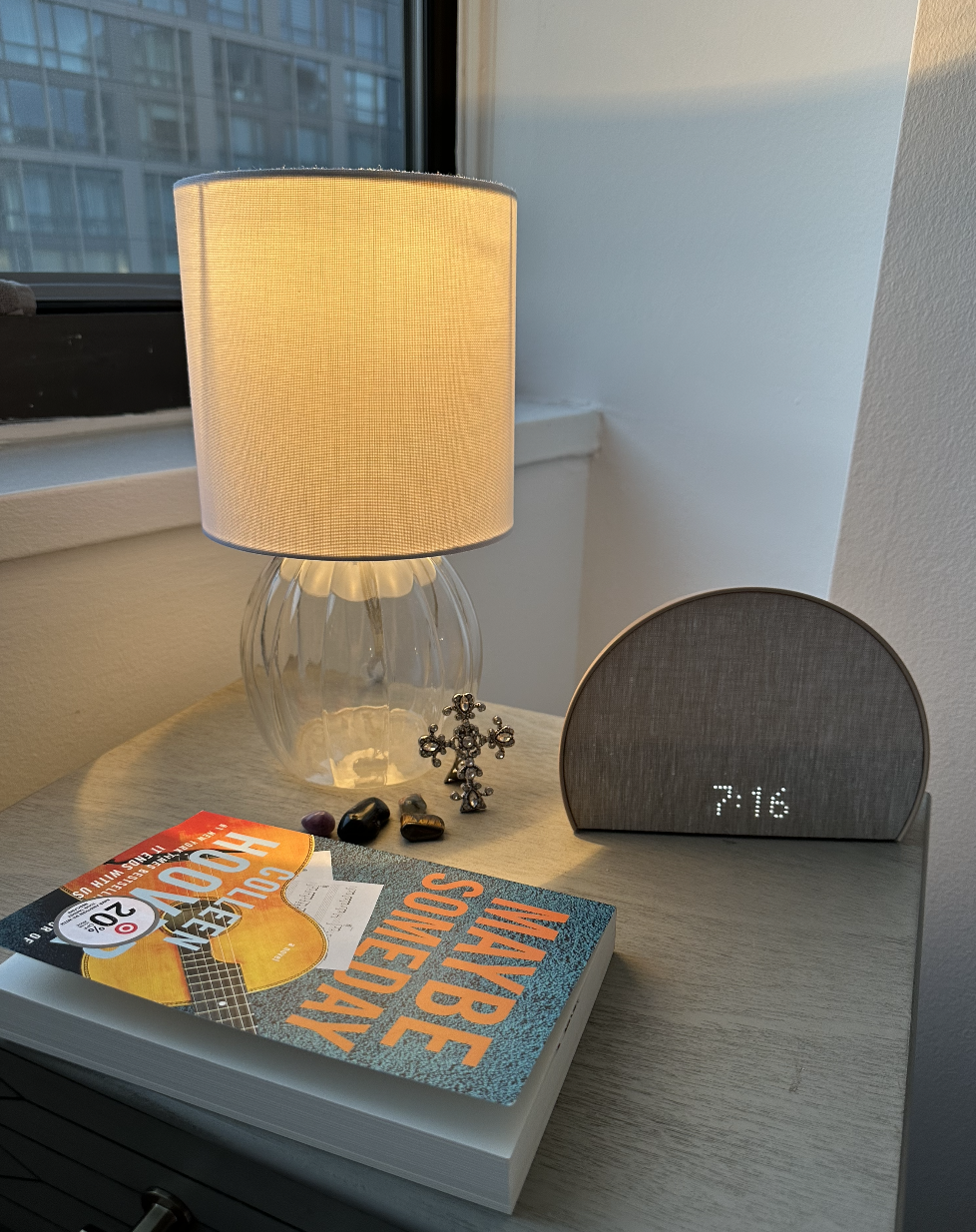 Table with lamp, alarm clock reading 9:16, and a book titled &quot;Maybe Someday&quot;. Decor includes earrings and stones