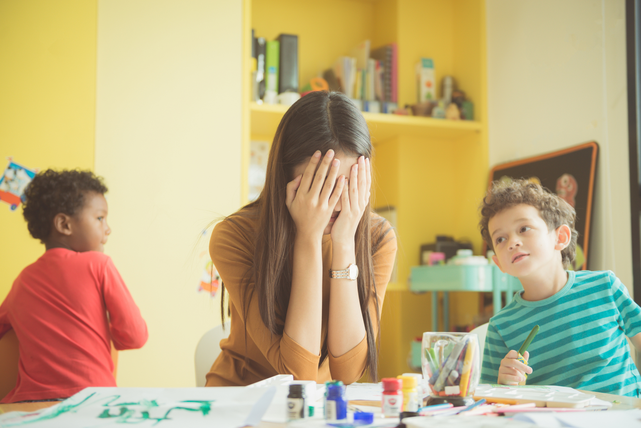 Adult covers face, two children with art supplies on table appear curious