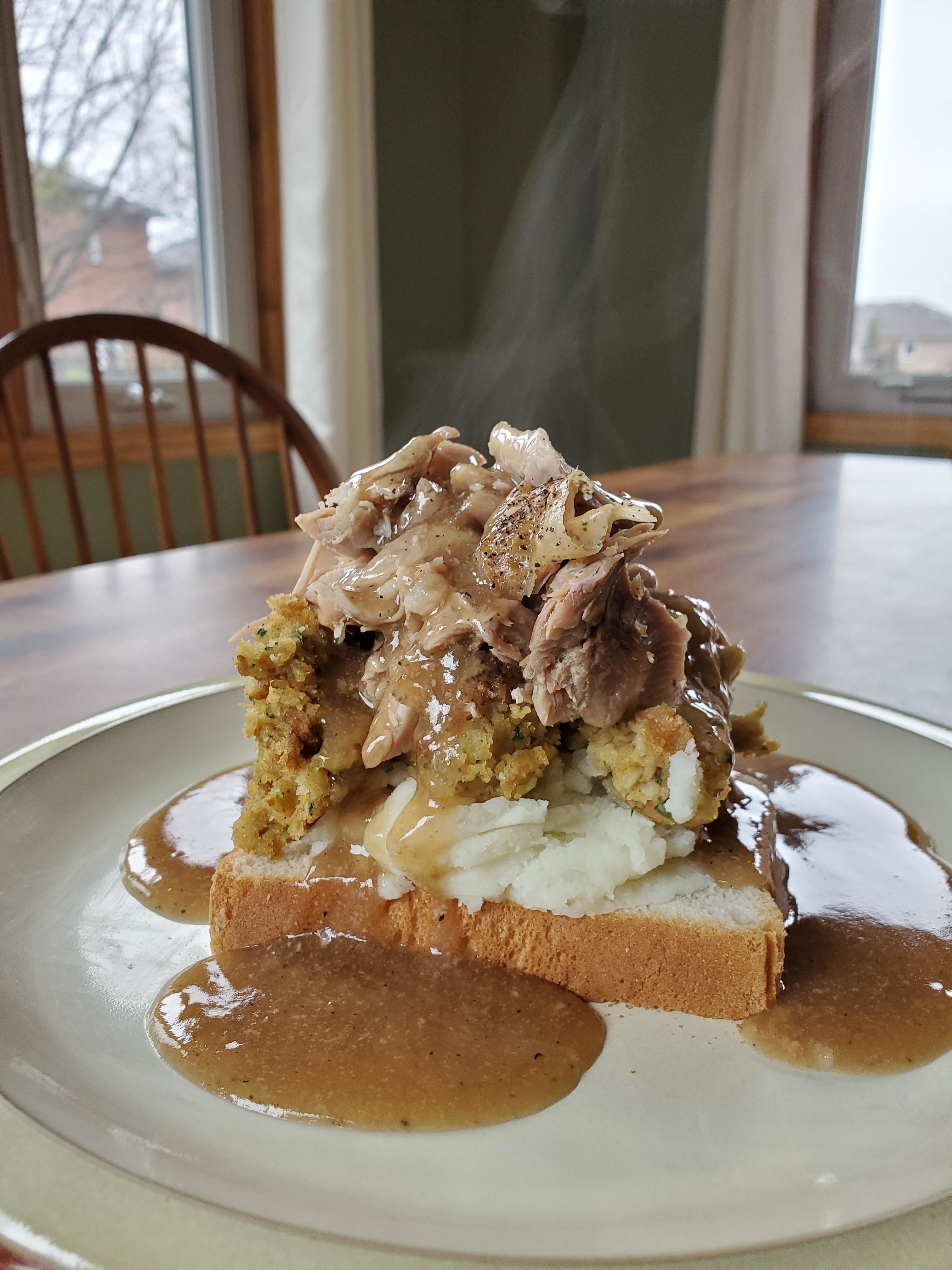 Open-faced turkey sandwich with stuffing, mashed potatoes, and gravy on a plate. Steam rises from the hot meal