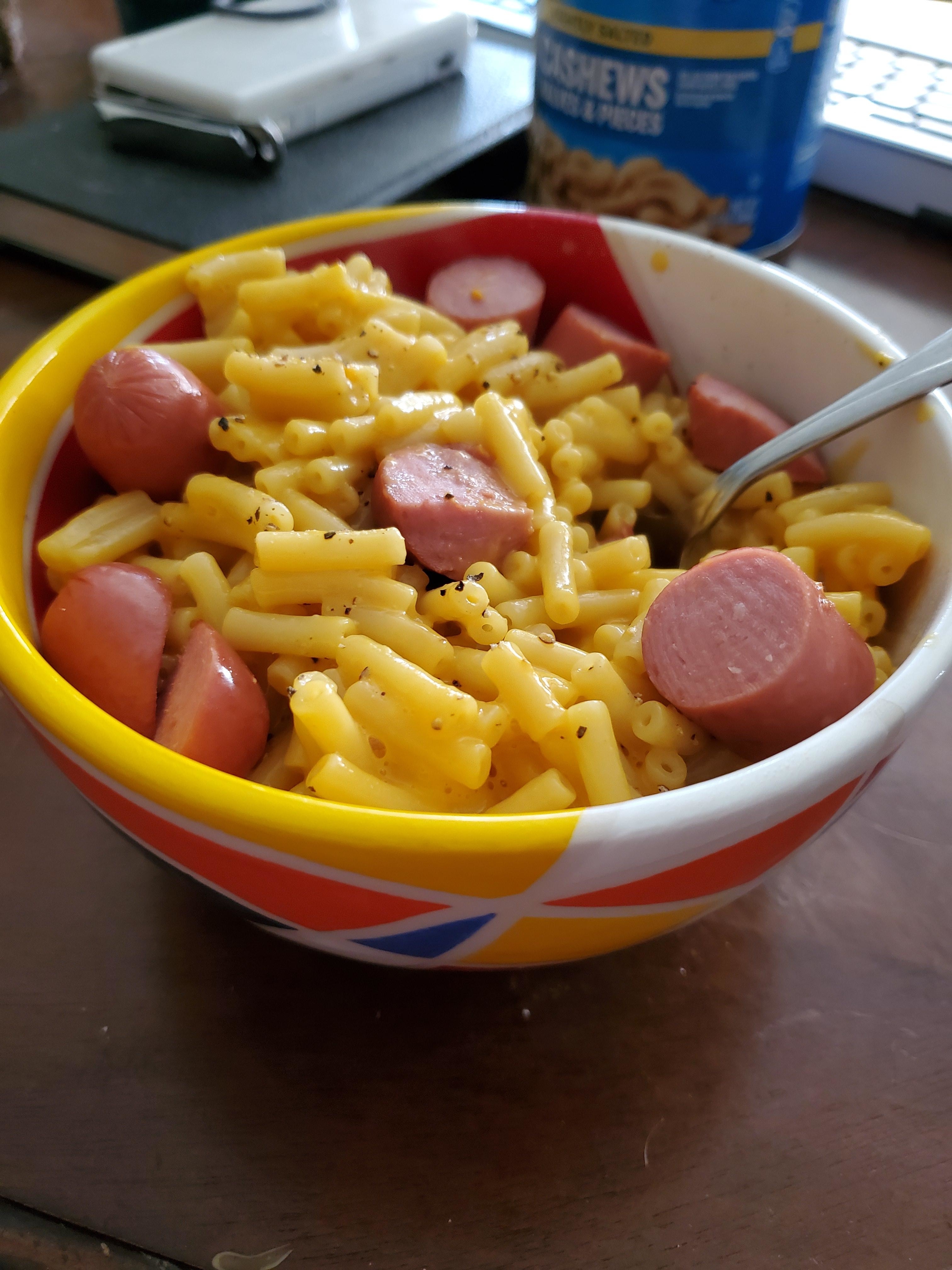 A bowl of macaroni and cheese with sliced hot dogs mixed in, on a table next to a can of cashews