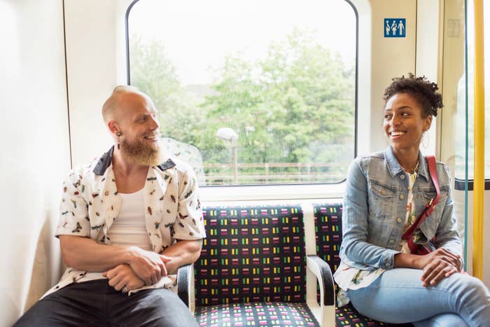 Two people smiling at each other while sitting on a train, one with a beard and printed shirt, the other with a denim jacket