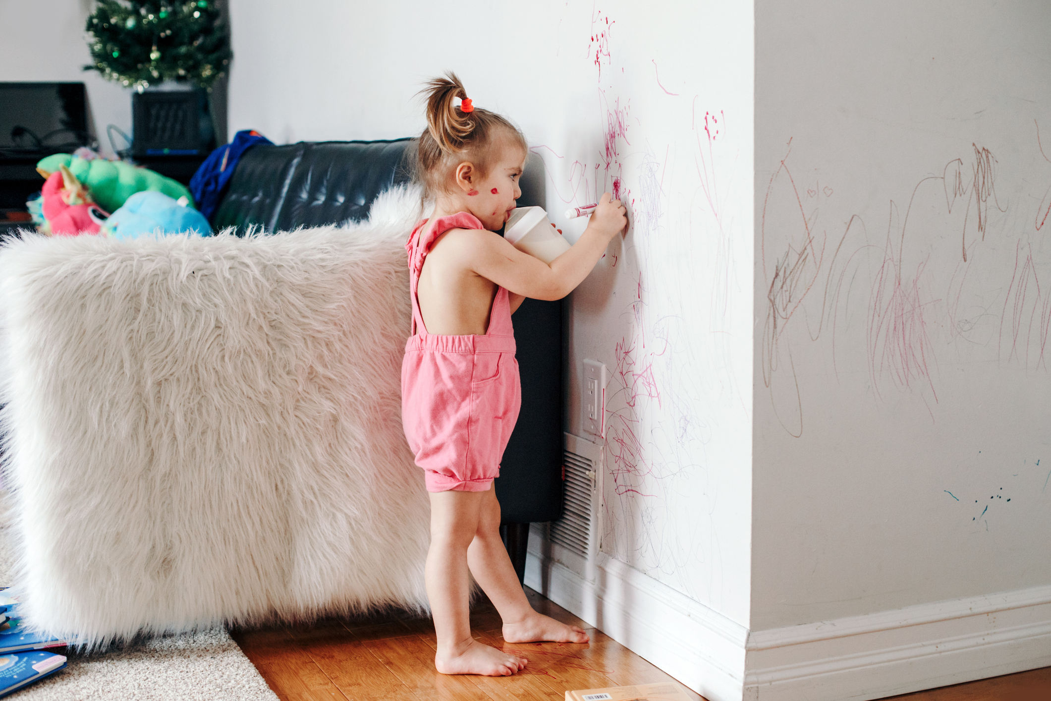 A toddler drawing on a wall next to a sofa and toys