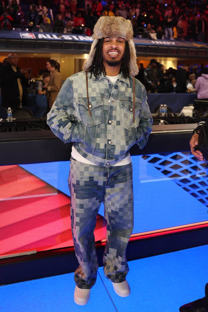 Man in camo-patterned denim outfit and fur hat smiling on a set with audience background