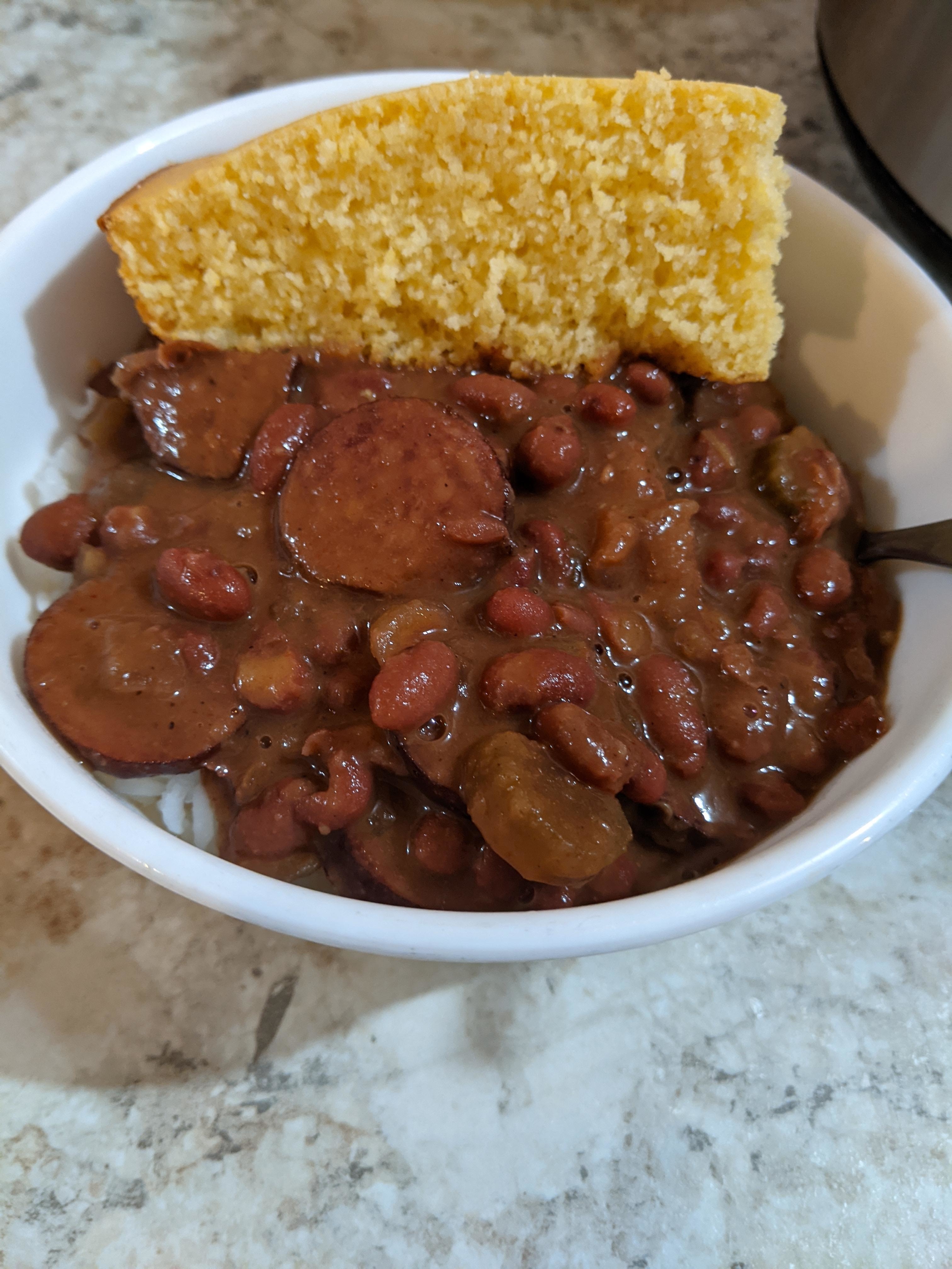A bowl of beans and slices of sausage, with a piece of cornbread on the side
