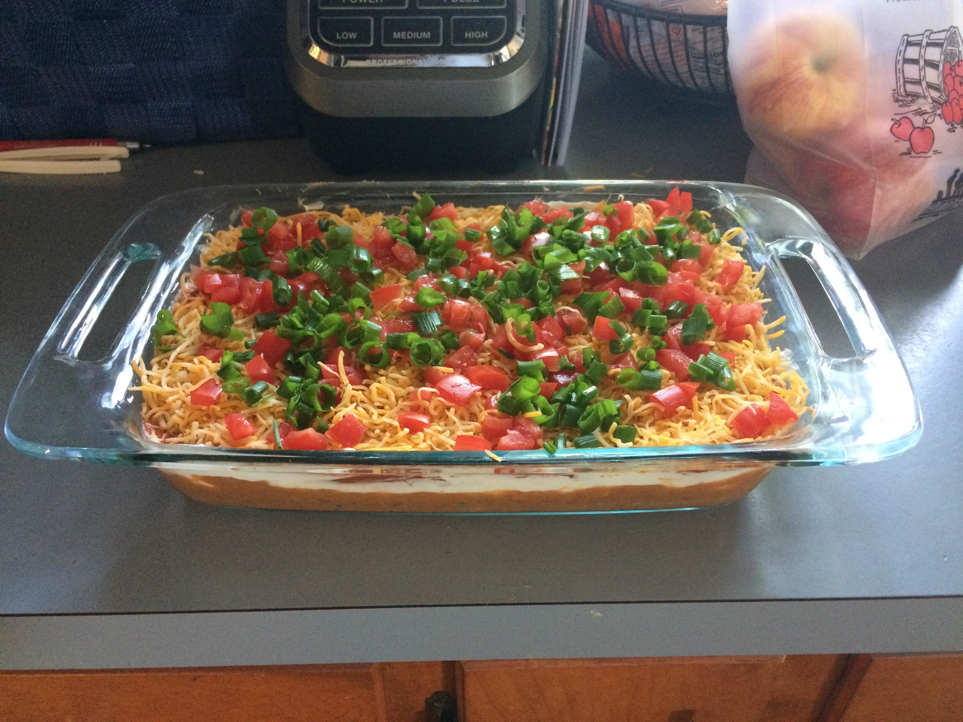 A glass baking dish filled with a layered taco dip topped with cheese, tomatoes, and green onions, on a kitchen counter