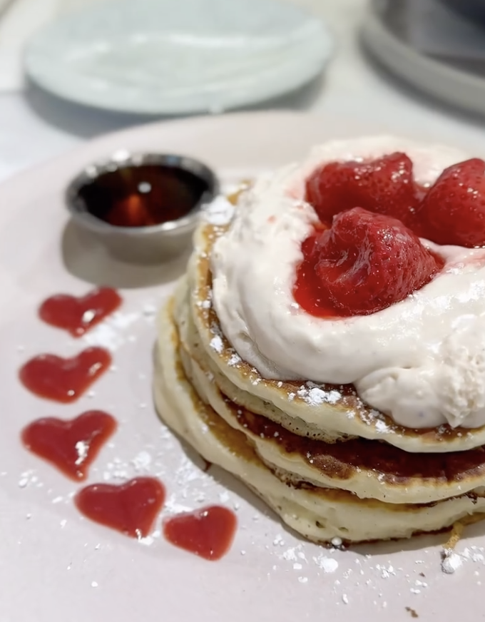 Stack of pancakes with cream and raspberries on top, garnished with heart-shaped syrup drops