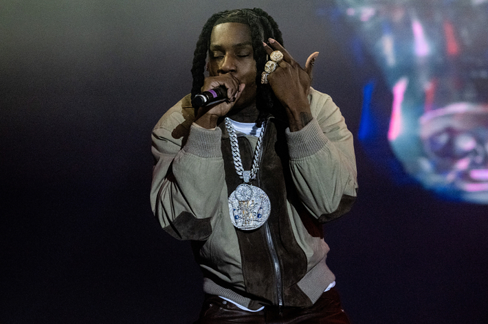 Rapper on stage performing, wearing heavy chain necklaces and rings, holding a microphone