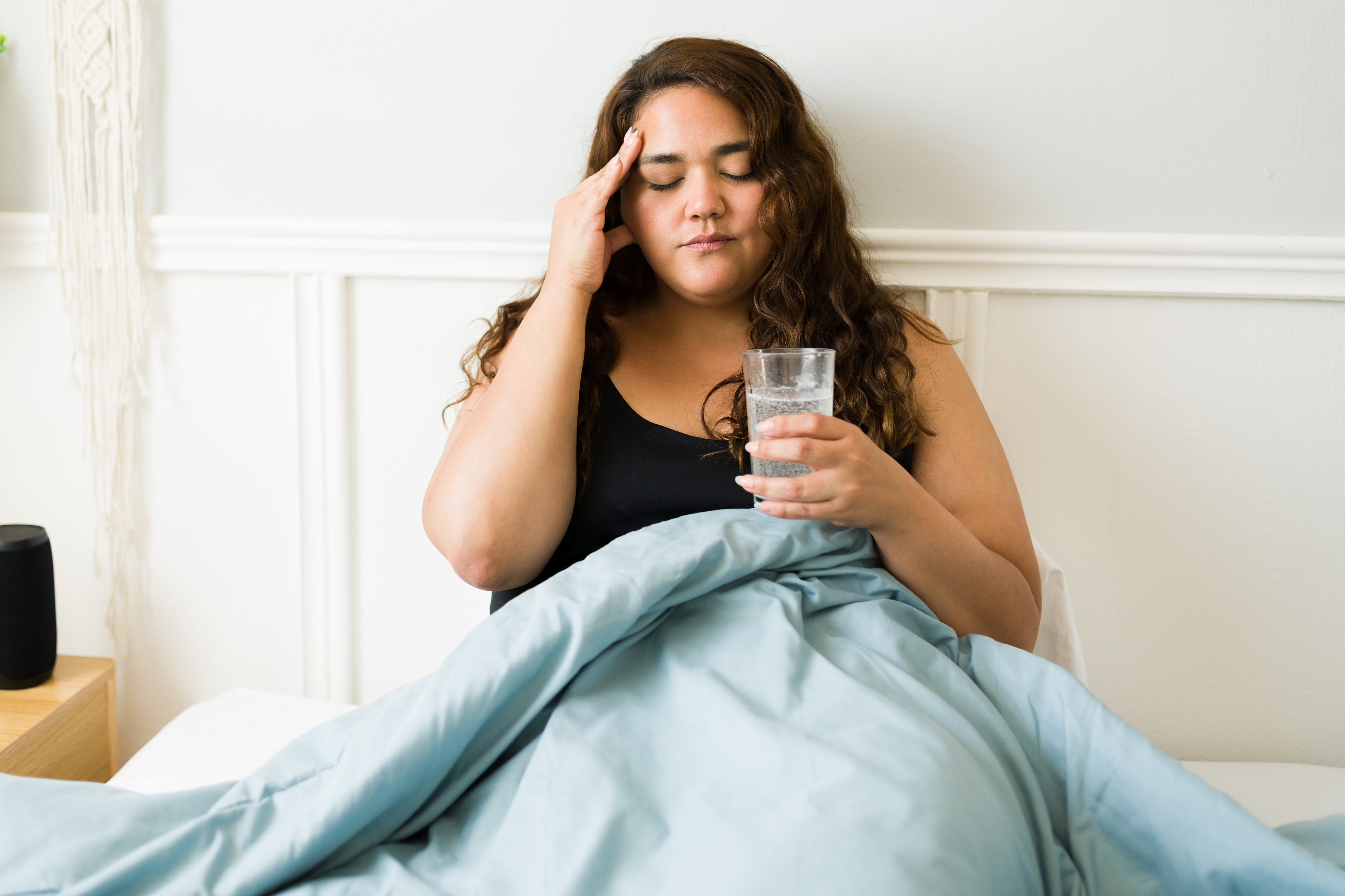Person in bed holding a glass of water and touching head, appearing unwell