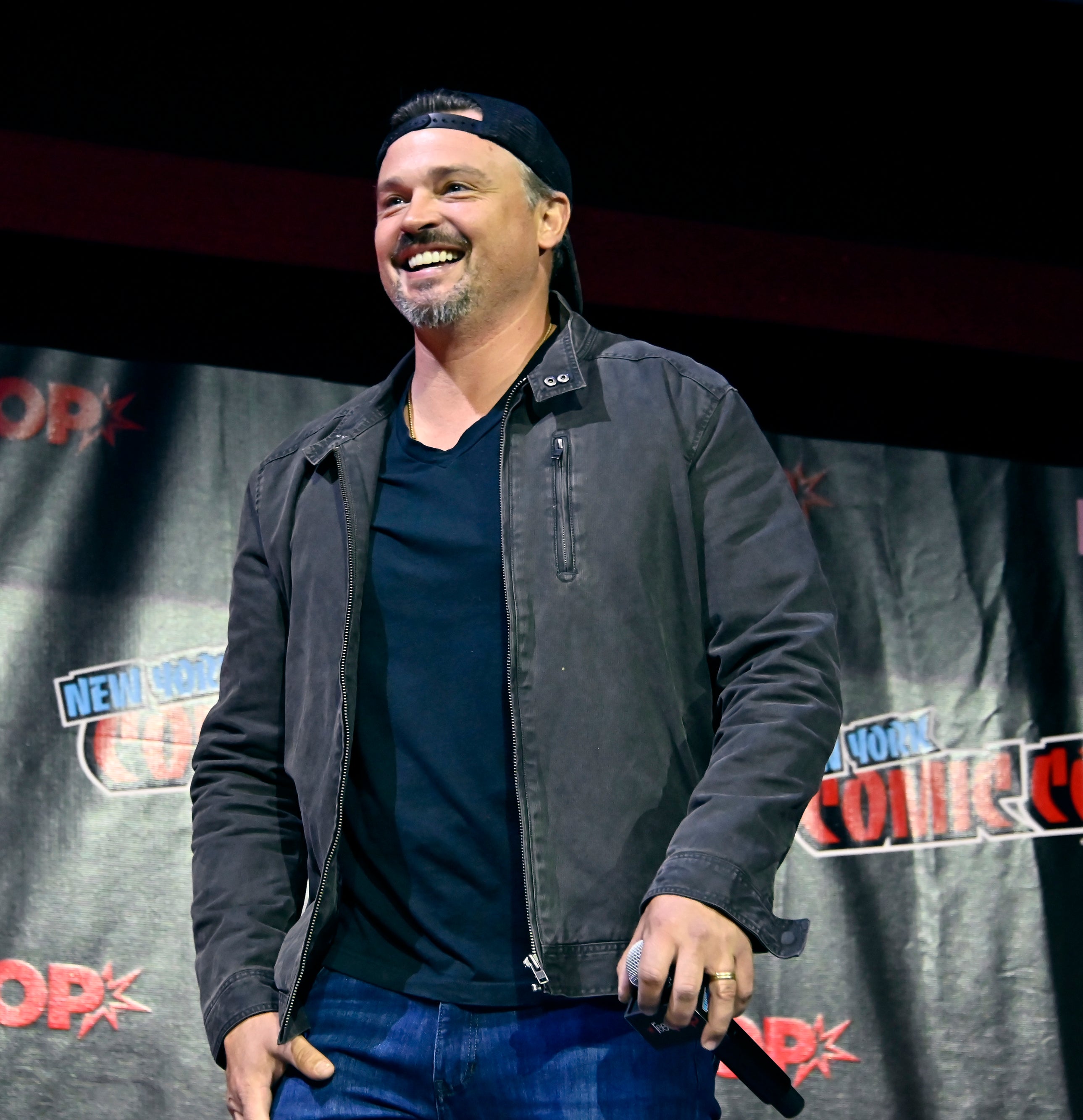 Man smiling on stage at New York Comic Con, wearing a headband, t-shirt, and jacket