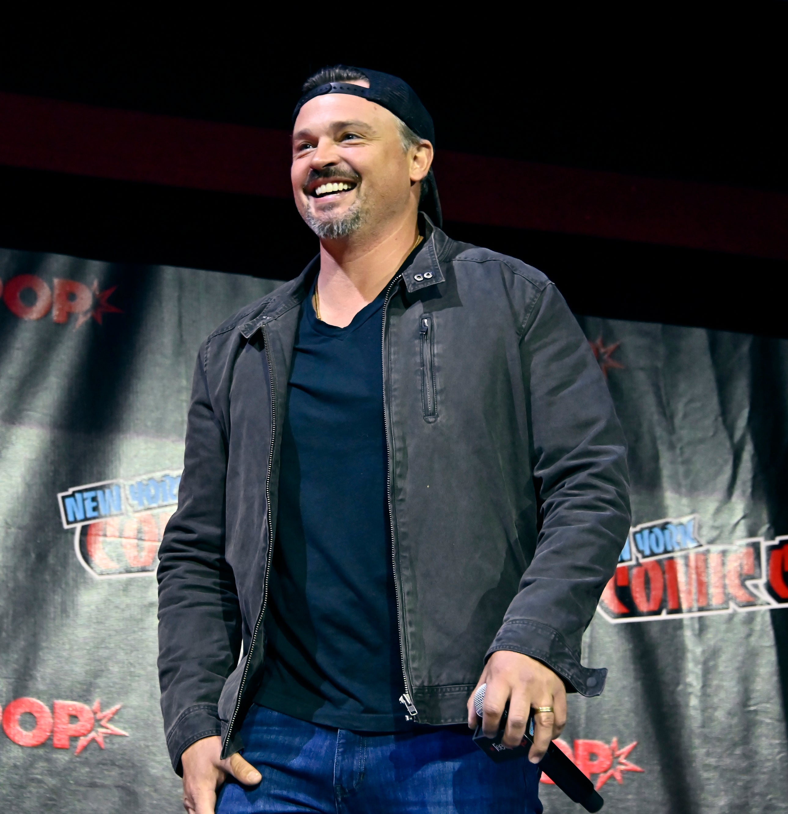 Man smiling on stage at New York Comic Con, wearing a headband, t-shirt, and jacket