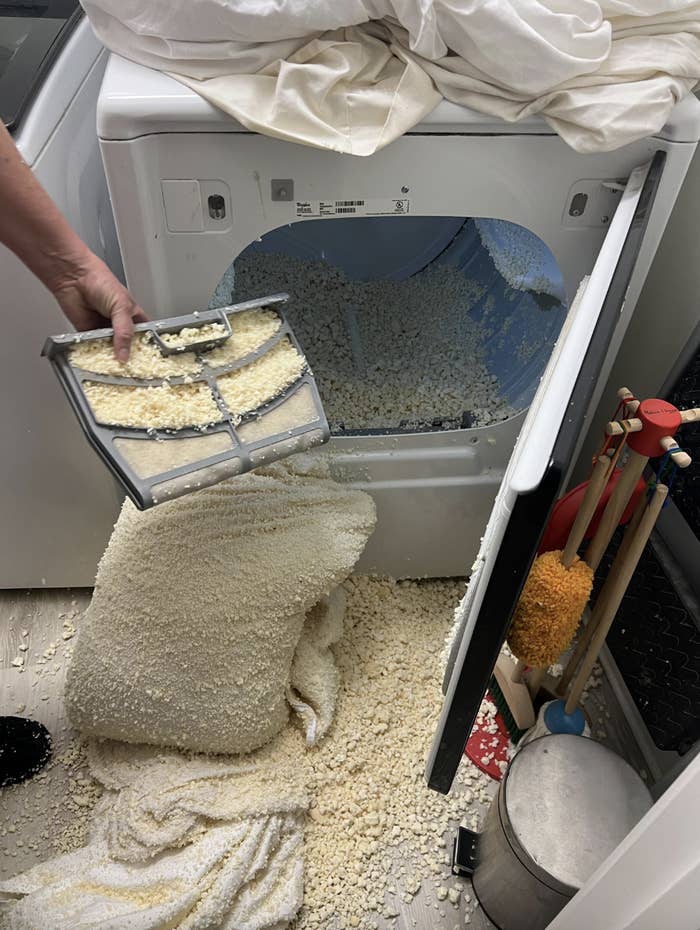 Washing machine overflows with detergent suds and spilled laundry beads, person holds the dispenser, cleaning tools nearby