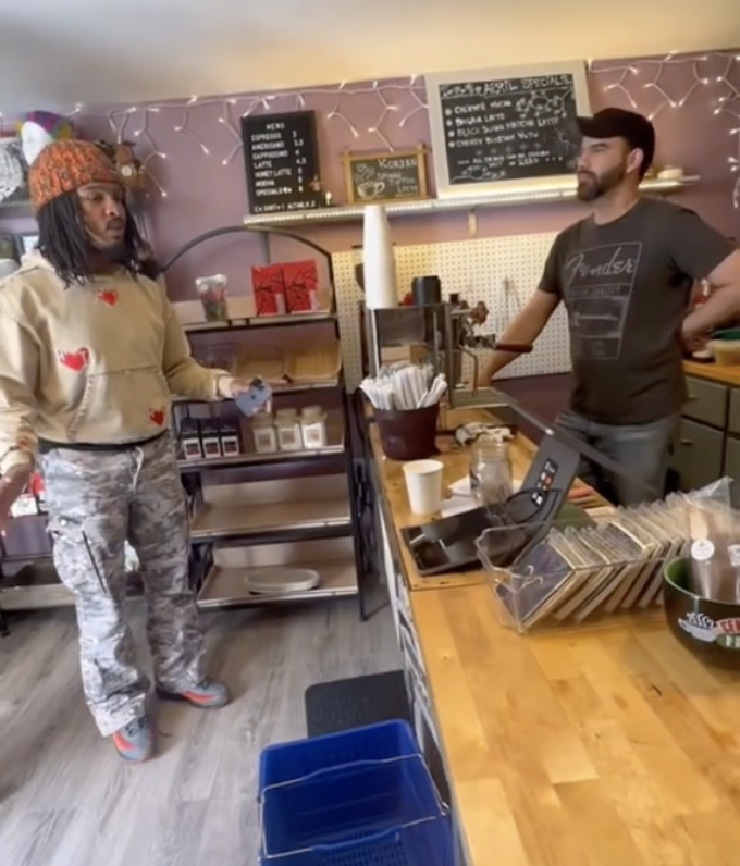 Two people stand in a casual café setting, one customer and one barista behind the counter. The customer gestures during a conversation