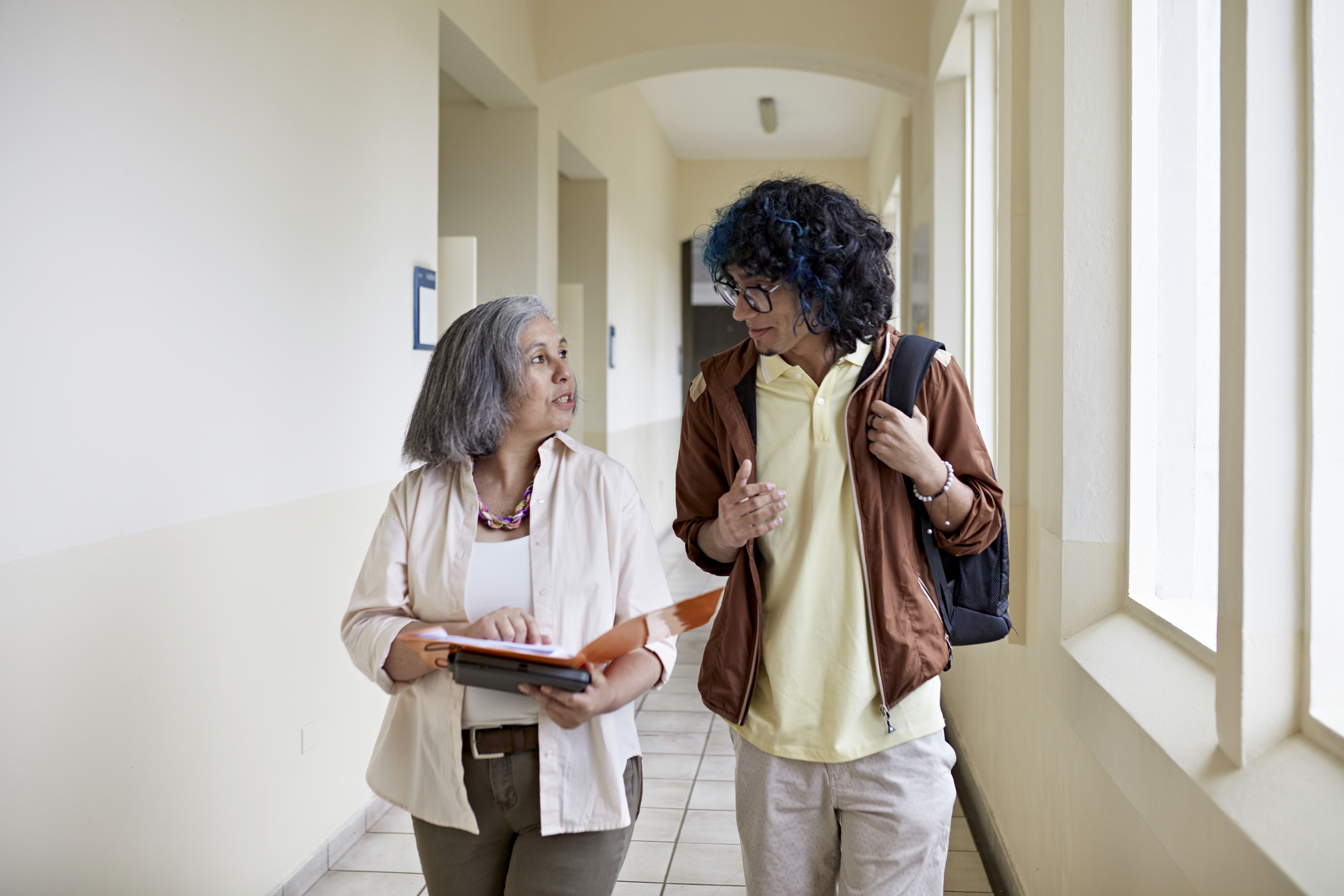 Two individuals conversing while walking through a hallway, one holding a tablet
