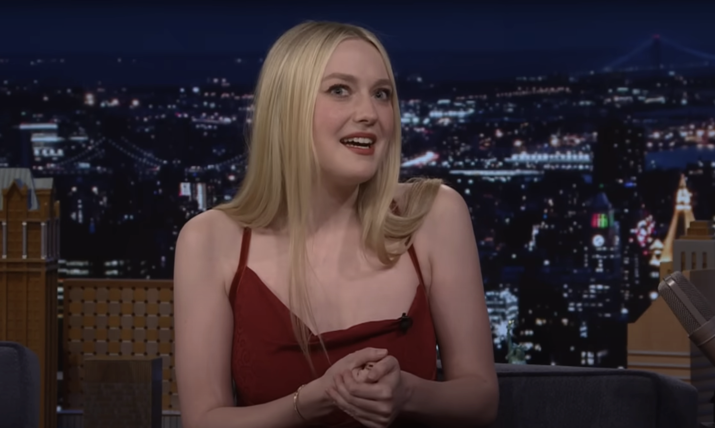 Woman in a red dress sits smiling on a talk show set with a cityscape background