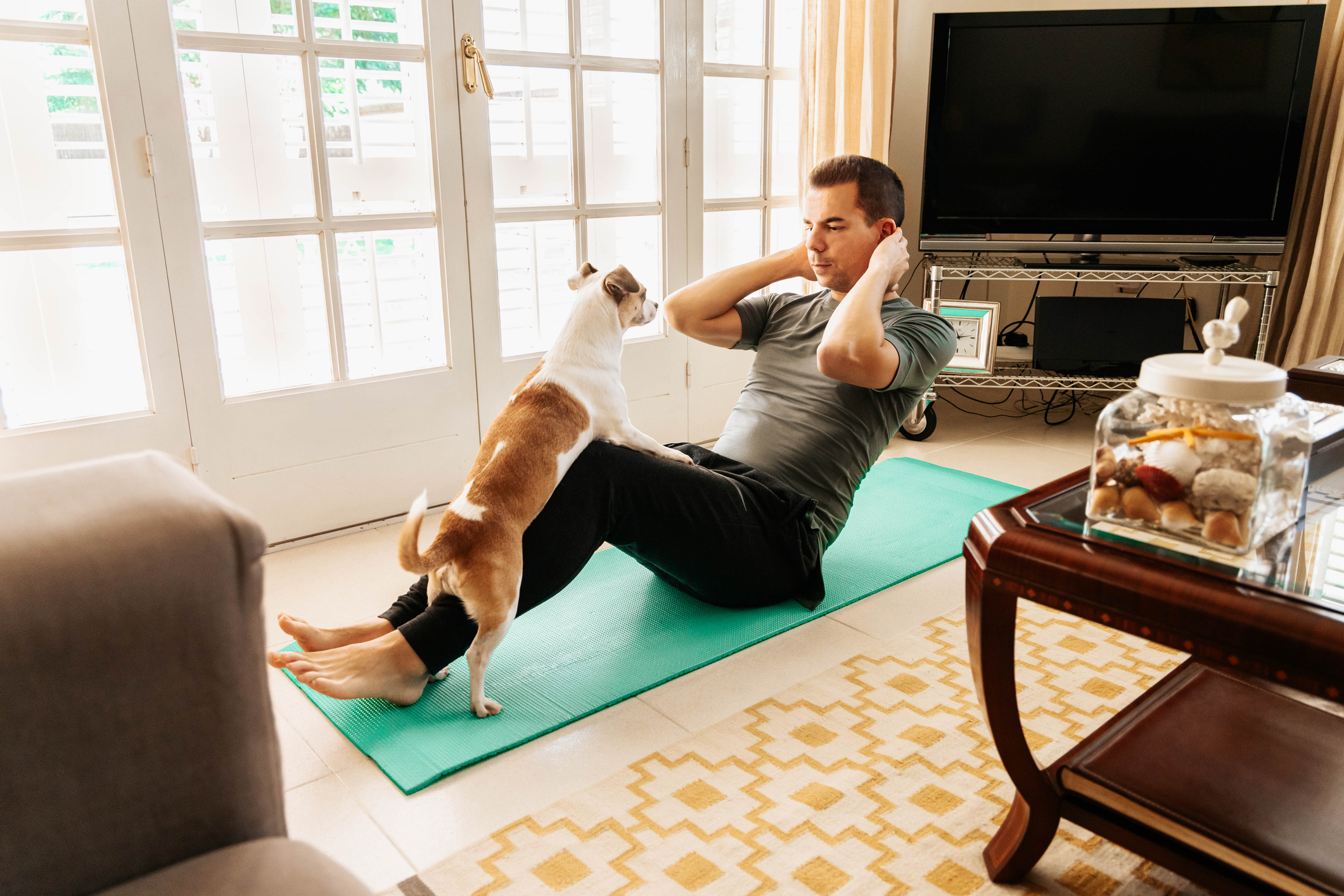 Man doing sit-ups at home with a dog beside him on a yoga mat, promoting fitness with pets