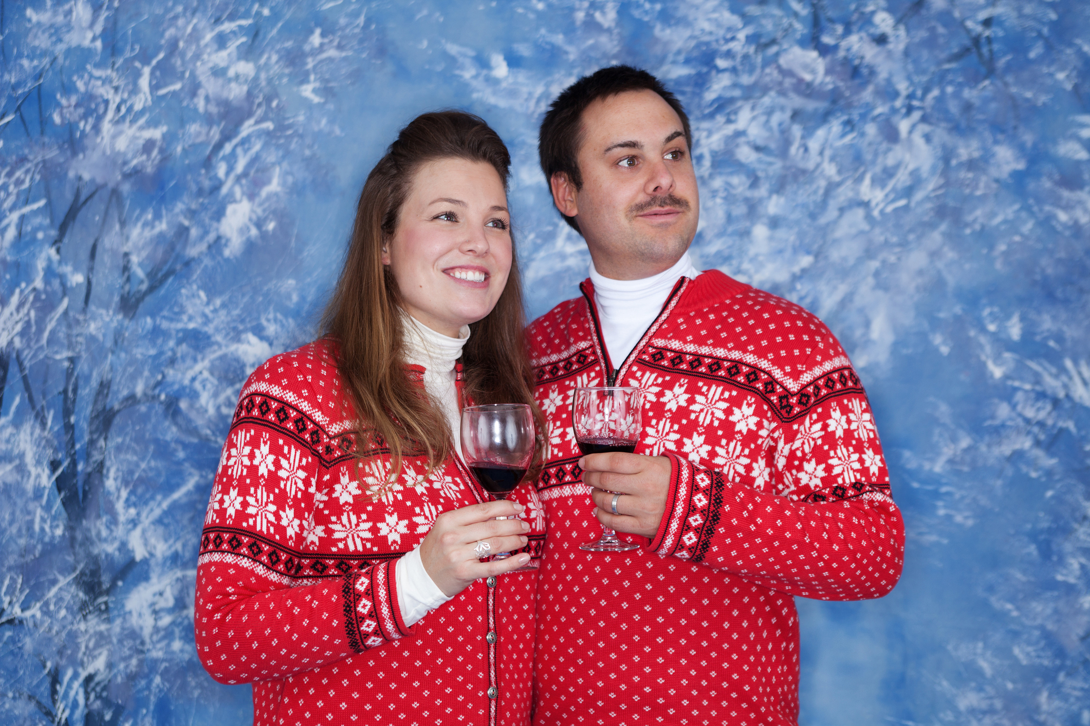 Two people in matching festive sweaters holding wine glasses, smiling, with a winter-themed backdrop