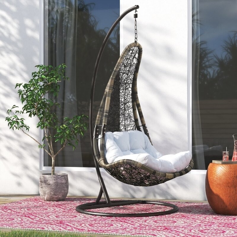 An empty hanging egg chair with cushions on a pink outdoor rug, beside a potted plant and a glass of drink